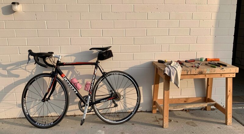 Two road bikes leaning against a plain painted brick wall beside a small wooden workbench with tools and a folded rag, afternoon light